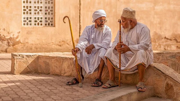 رجلان مسنّان بالدشداشة البيضاء والعمامة، يجلسان على دكّة حجرية أمام جدار طيني بنوافذ مشربية، يتّكئ كلّ منهما على عصاه، ويتحادثان في سكينة تامة.